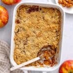 Apple crumble in the baking dish with serving removed to a small plate nearby, serving spoon dipped inside, a few apples around.