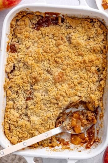 Apple crumble in the baking dish with serving removed to a small plate nearby, serving spoon dipped inside, a few apples around.