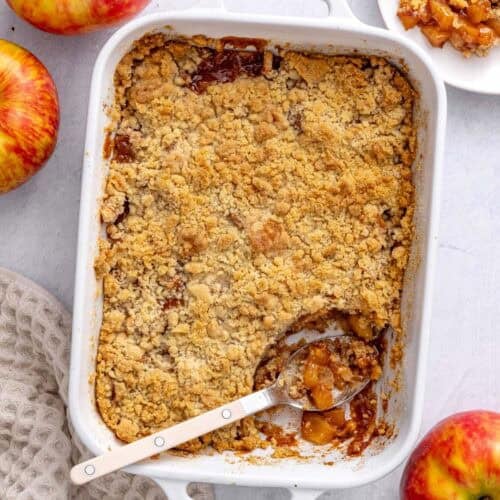 Apple crumble in the baking dish with serving removed to a small plate nearby, serving spoon dipped inside, a few apples around.