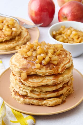 Two plates of pancakes with apple compote on top with an extra small dish of more compote and some whole apples around.