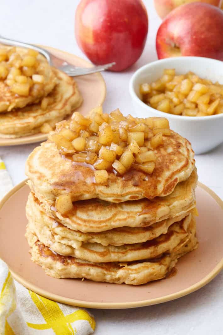 Two plates of pancakes with apple compote on top with an extra small dish of more compote and some whole apples around.