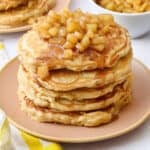 Two plates of pancakes with apple compote on top with an extra small dish of more compote and some whole apples around.