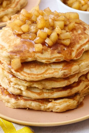 Two plates of pancakes with apple compote on top with an extra small dish of more compote and some whole apples around.