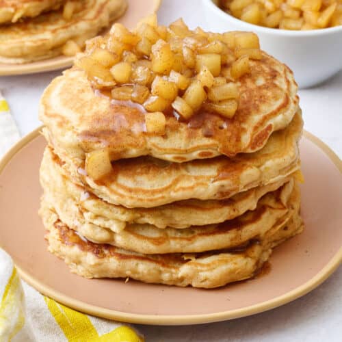 Two plates of pancakes with apple compote on top with an extra small dish of more compote and some whole apples around.