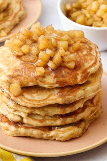 Two plates of pancakes with apple compote on top with an extra small dish of more compote and some whole apples around.