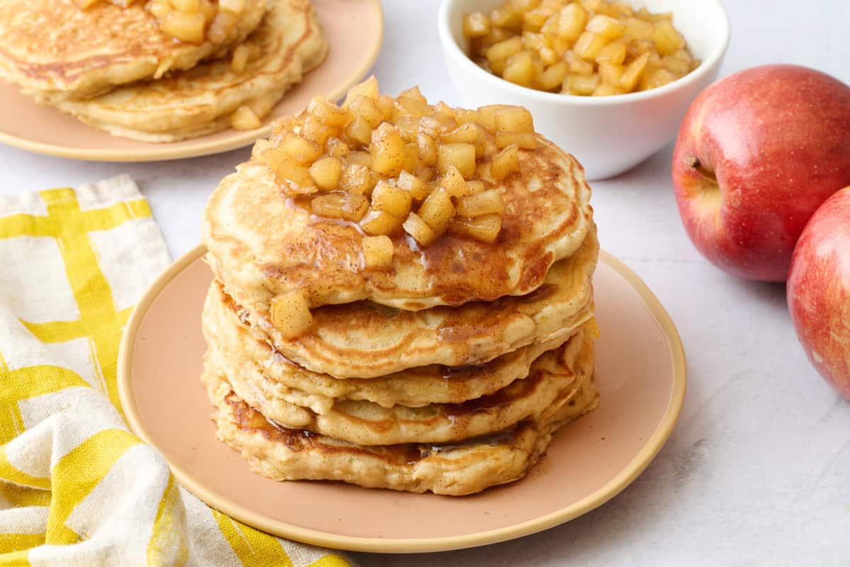 Two plates of pancakes with apple compote on top with an extra small dish of more compote and some whole apples around.