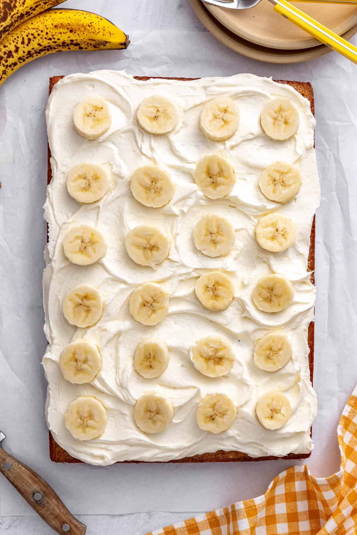 Iced cake in the cake pan with bananas topping it and whole bananas nearby,