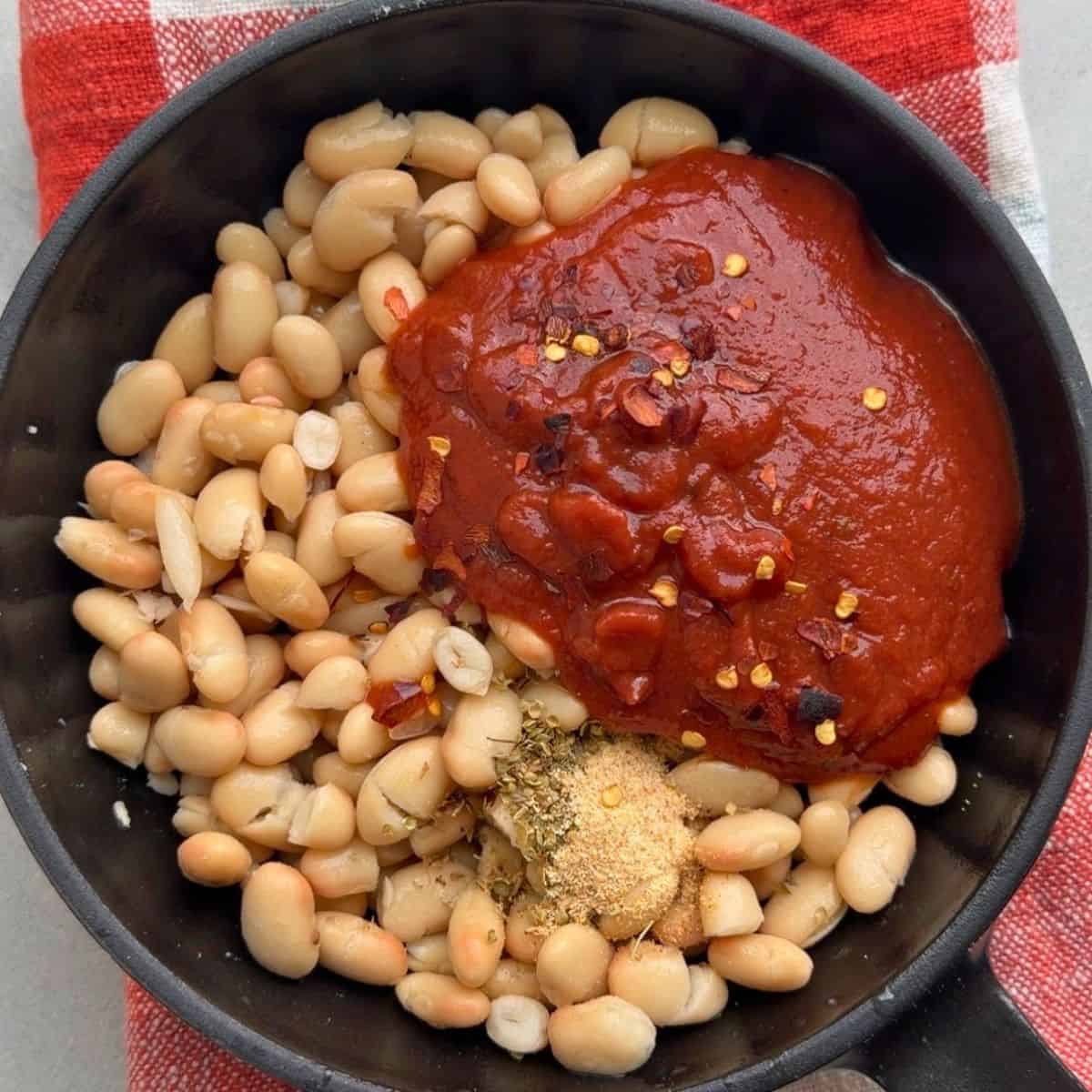 Marinara sauce and seasonings added on top of beans in a skillet before combining.