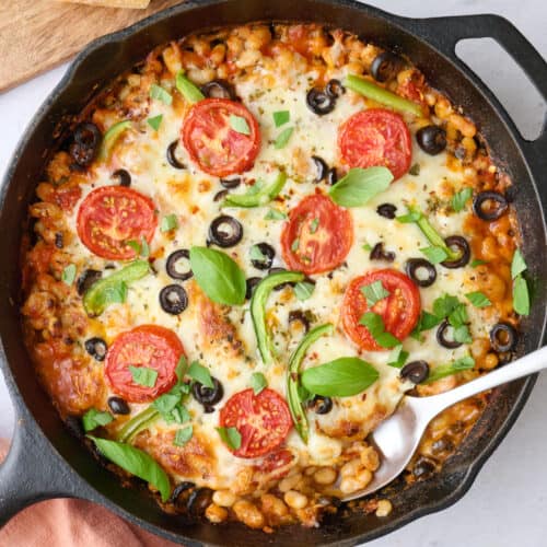 Pizza beans garnished with fresh basil with crusty bread nearby, serving removed with spoon dipped inside.
