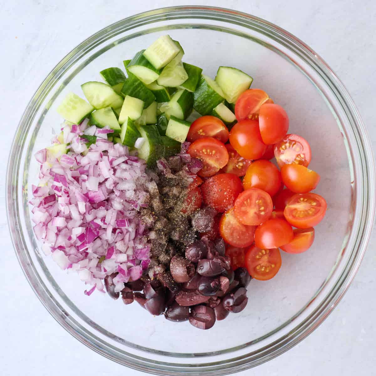 Tomatoes, cucumbers, olives, onions, oil and seasonings in a bowl before tossing.
