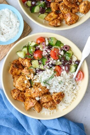 Two Greek chicken bowls served over rice with chicken, veggie salad, and tzatziki segmented on top, garnished with feta and fresh dill with a dish of more tzatziki nearby.