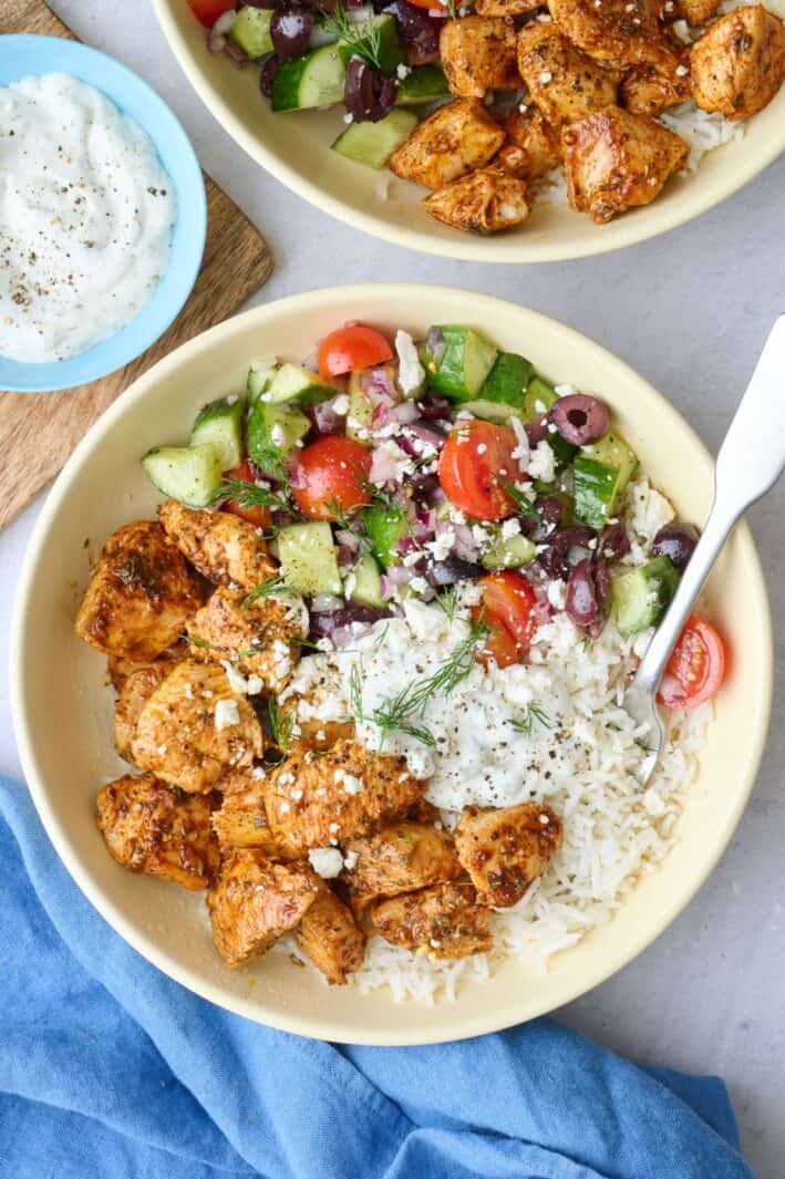 Two Greek chicken bowls served over rice with chicken, veggie salad, and tzatziki segmented on top, garnished with feta and fresh dill with a dish of more tzatziki nearby.