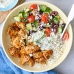 A Greek chicken bowl served over rice with chicken, veggie salad, and tzatziki segmented on top, garnished with feta and fresh dill with a dish of more tzatziki nearby.