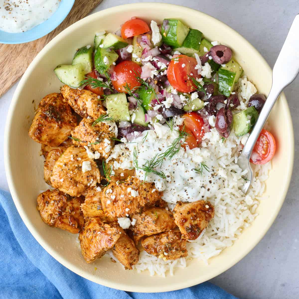 A Greek chicken bowl served over rice with chicken, veggie salad, and tzatziki segmented on top, garnished with feta and fresh dill with a dish of more tzatziki nearby.