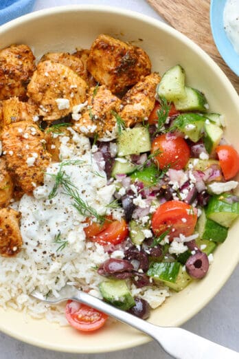 Two Greek chicken bowls served over rice with chicken, veggie salad, and tzatziki segmented on top, garnished with feta and fresh dill with a dish of more tzatziki nearby.