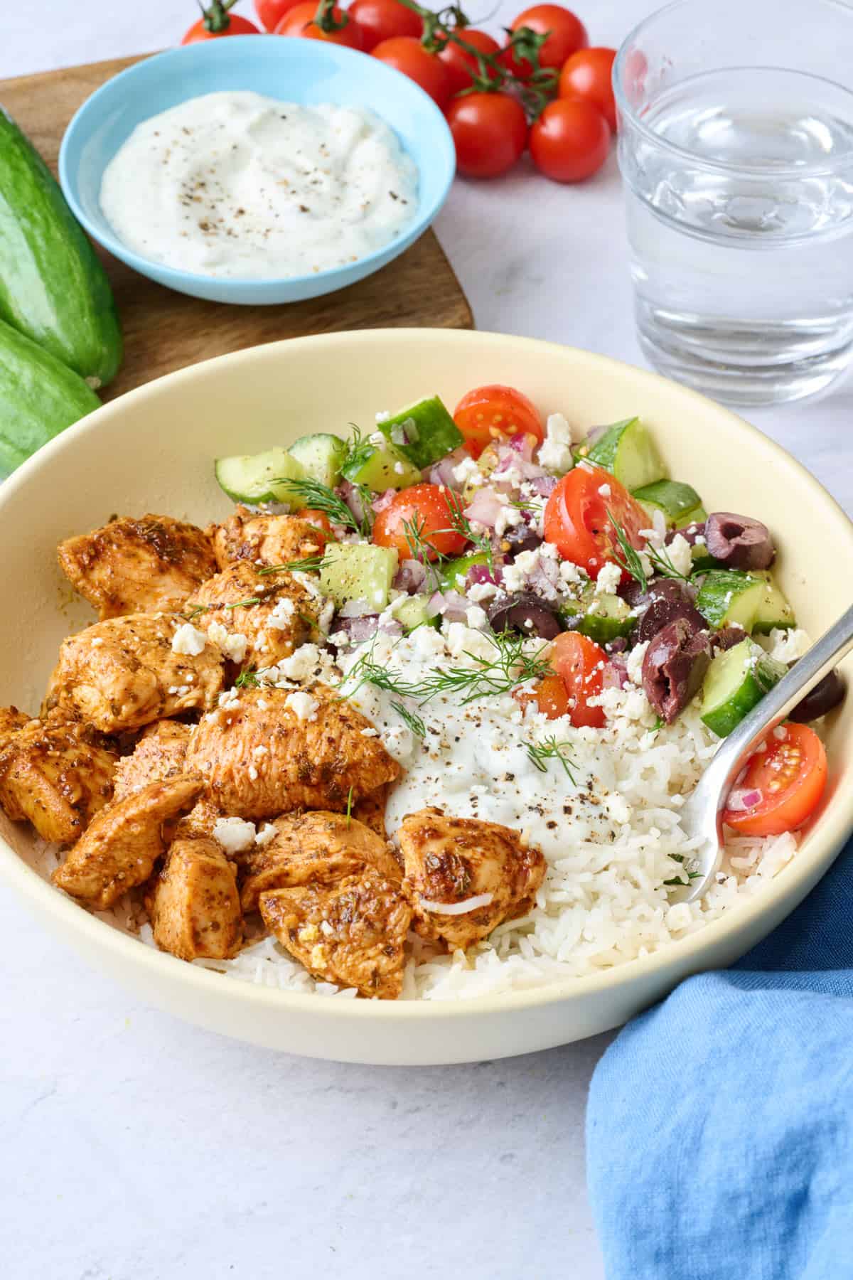 A Greek chicken bowl served over rice with chicken, veggie salad, and tzatziki segmented on top, garnished with feta and fresh dill with a dish of more tzatziki nearby.