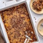 Pecan pie cobbler in a baking dish with a serving removed to a small plate nearby, serving spoon dipped inside baking dish with the gooey sauce visible.