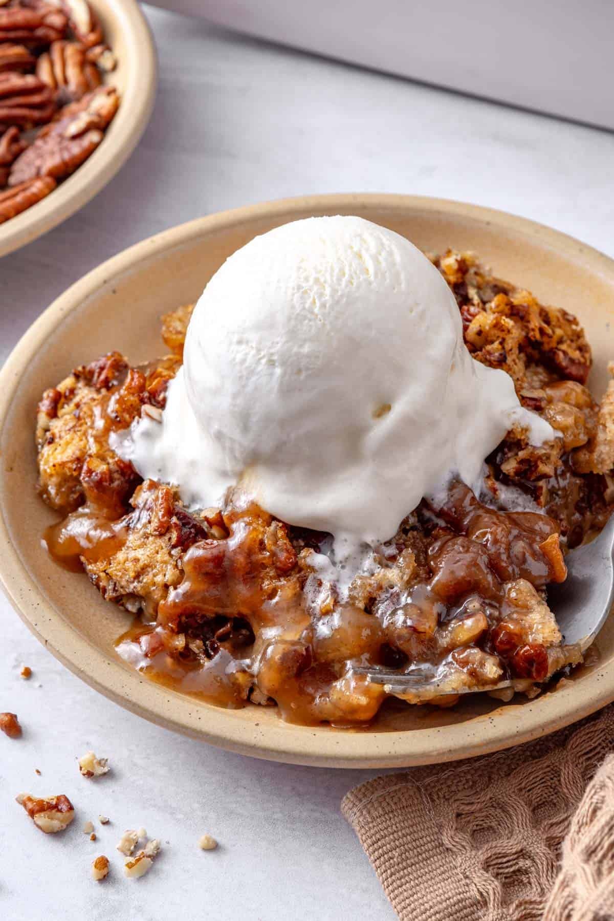 Serving of cobbler on a plate with ice cream on top, baking dish nearby.