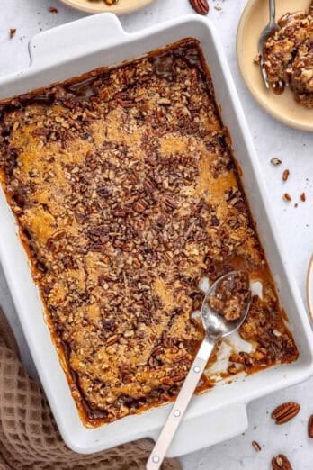 Pecan pie cobbler in a baking dish with a serving removed to a small plate nearby, serving spoon dipped inside baking dish with the gooey sauce visible.