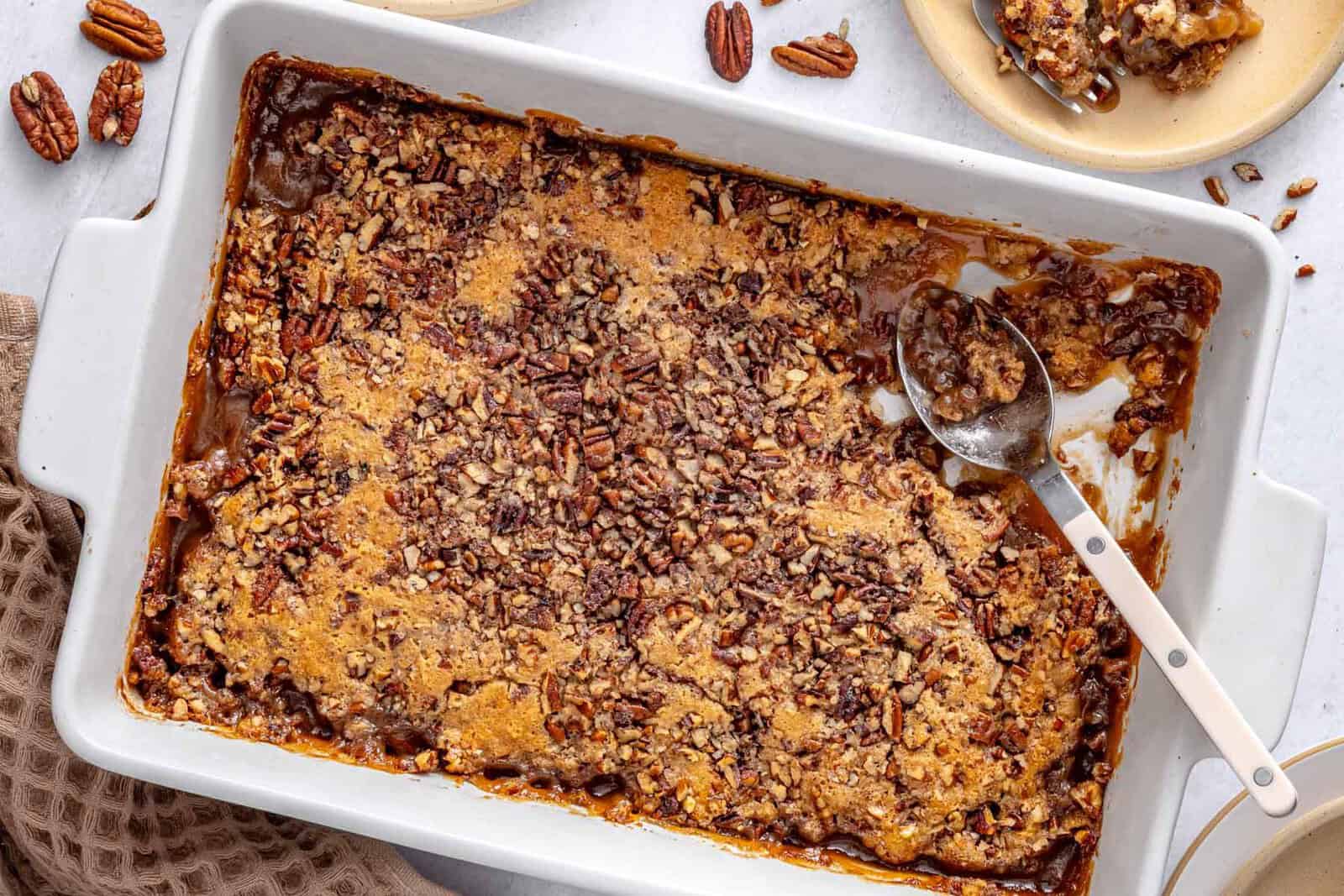 Pecan pie cobbler in a baking dish with a serving removed to a small plate nearby, serving spoon dipped inside baking dish with the gooey sauce visible.