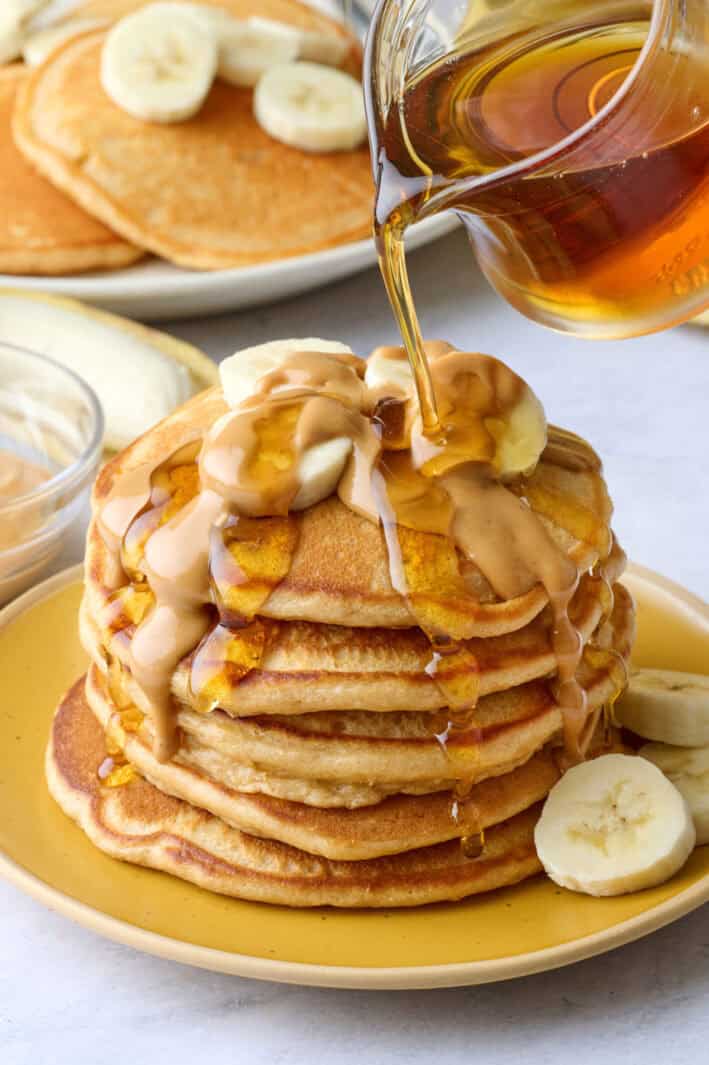 Peanut butter pancakes stacked on a plate with sliced bananas on top, melted, drippy peanut butter poured over and maple syrup being poured lightly on top.