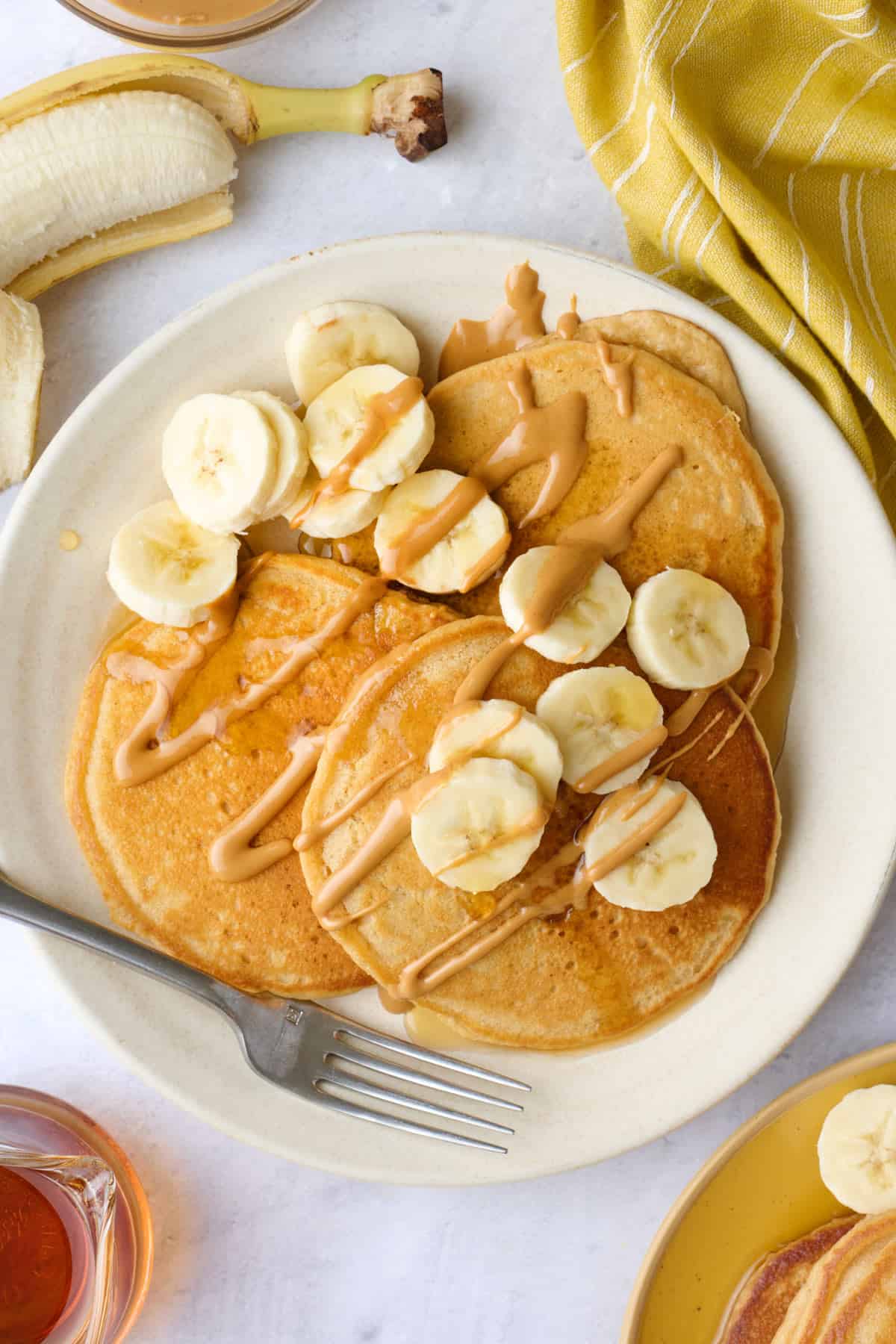 Three pancakes on a plate with peanut butter drizzle and banana slices on top and more pancakes and ingredients nearby.