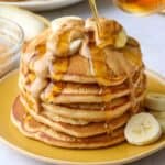 Peanut butter pancakes stacked on a plate with sliced bananas on top, melted, drippy peanut butter poured over and maple syrup being poured lightly on top.