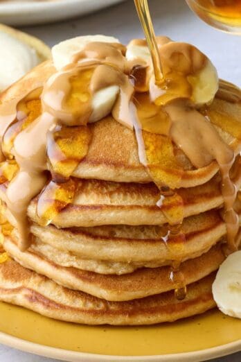 Peanut butter pancakes stacked on a plate with sliced bananas on top, melted, drippy peanut butter poured over and maple syrup being poured lightly on top.