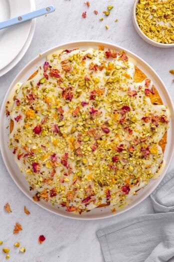 Persian love cake topped with glaze, pistachios, rose petals and orange zest, with more ingredients nearby.