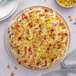 Persian love cake topped with glaze, pistachios, rose petals and orange zest, with more ingredients nearby.