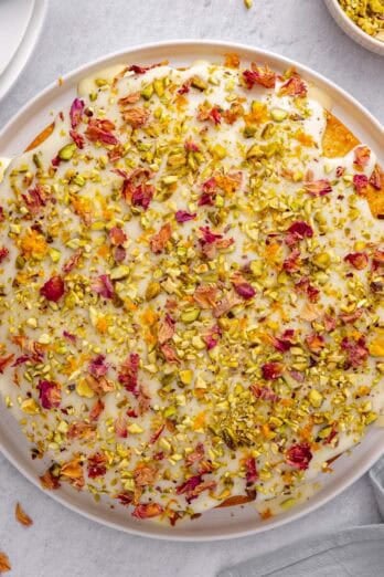 Persian love cake topped with glaze, pistachios, rose petals and orange zest, with more ingredients nearby.