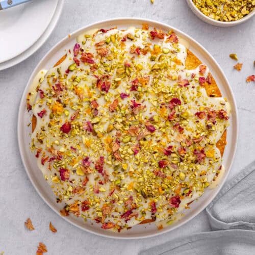 Persian love cake topped with glaze, pistachios, rose petals and orange zest, with more ingredients nearby.