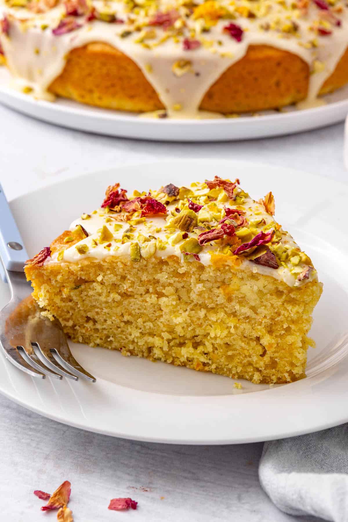 A slice of cake on a small plate with a fork and the whole cake in background.
