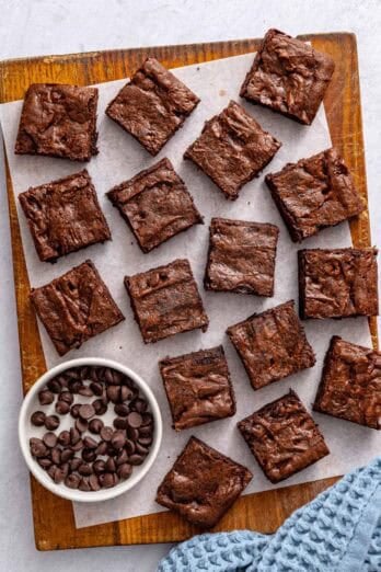 Chocolate brownies cut into 16 squares on a cutting board with a small dish of extra chocolate chips nearby.