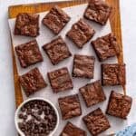 Chocolate brownies cut into 16 squares on a cutting board with a small dish of extra chocolate chips nearby.