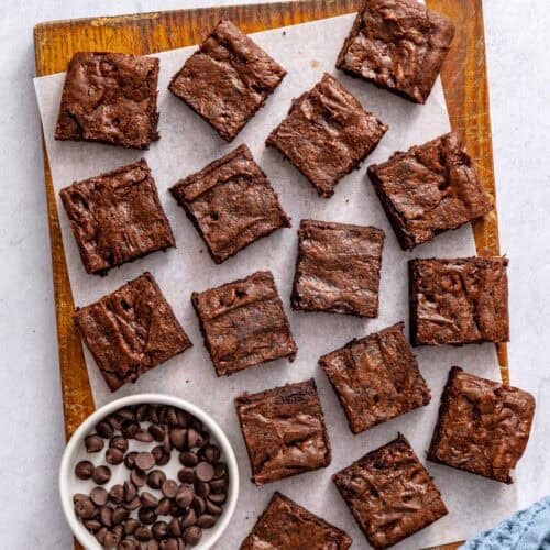 Chocolate brownies cut into 16 squares on a cutting board with a small dish of extra chocolate chips nearby.