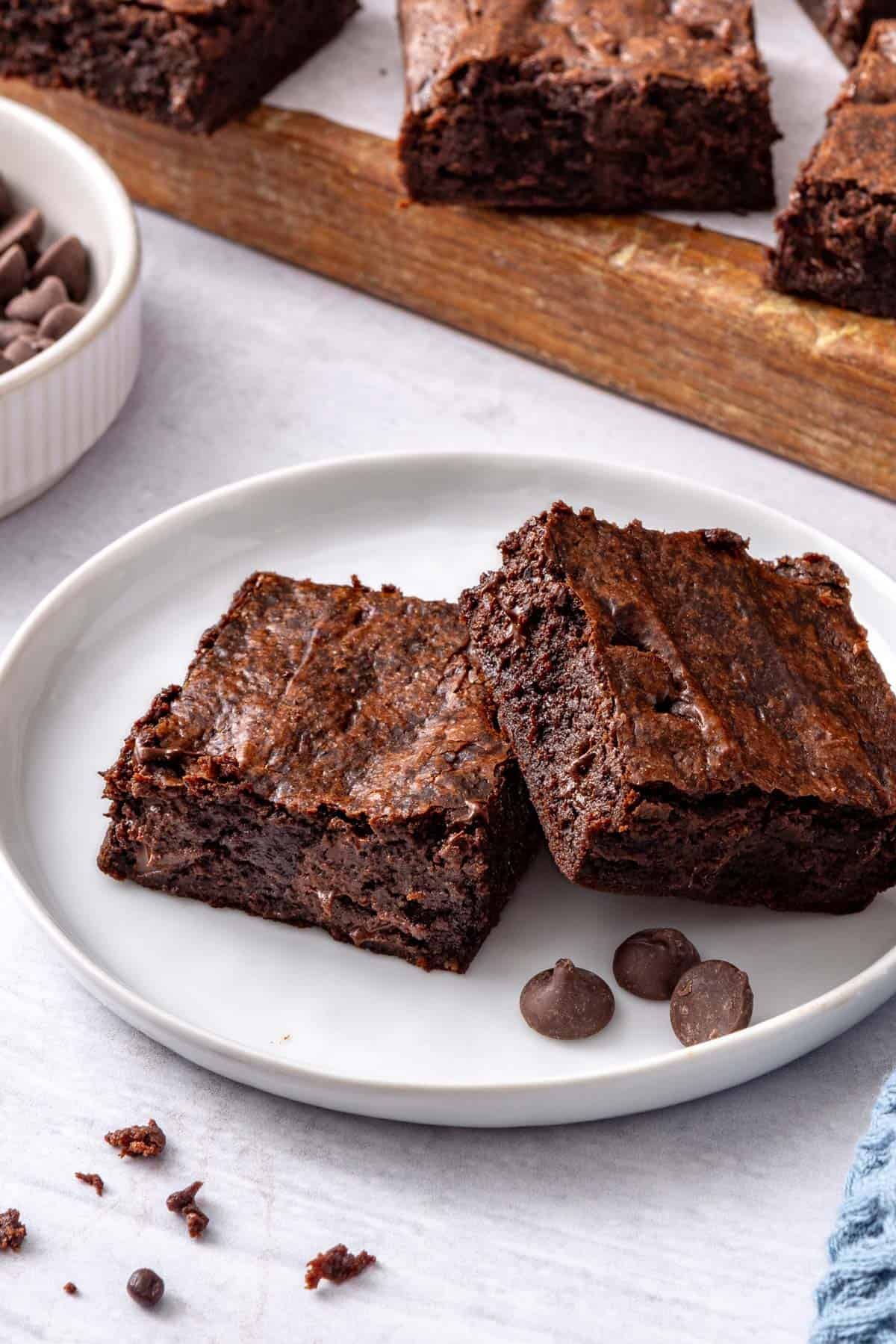 Two brownie squares layered on a small dessert plate with a board of more brownies and dish of chocolate chips nearby.